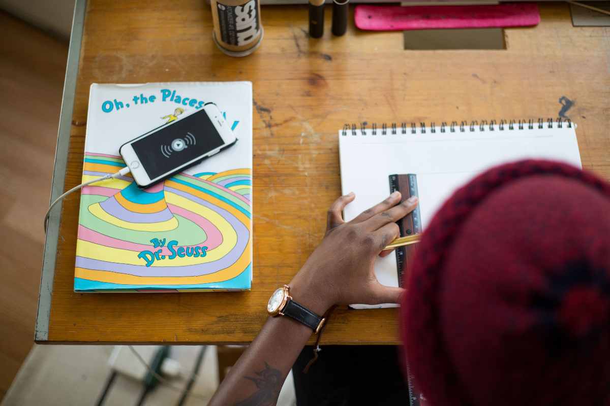 como-se-concentrar-nos-estudos-Pessoa negra faz anotação com uma régua em um caderno, enquanto o celular carrega ao lado em cima da mesa.